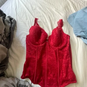 Röd spetskorsett med breda axelband - Snygg röd korsett med spetsdetaljer och breda axelband. Kuporna är vadderade och ger bra stöd, och baksidan har flera hyskor för justerbar passform. Perfekt för att lyfta vilken outfit som helst med en touch av edge.