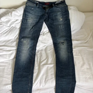 Jack & Jones skinny jeans blå 29/32 - Snygga blå skinny jeans från Jack & Jones, modell Liam. Jeansen har slitna detaljer och ljusa partier för en cool, använd look. Klassisk femficksmodell med dragkedjegylf och bruna sömmar. Perfekta för dig som gillar en smal passform.