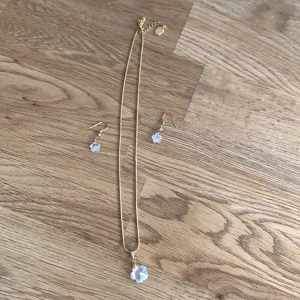 Guldigt smyckeset med blå blommor - Snyggt set med halsband och örhängen i guldton. Både halsbandet och örhängena har små blå blommor i glas eller kristall som hänger som berlocker. Perfekt för dig som gillar söta detaljer och vill sticka ut lite extra.