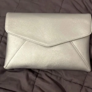 Silver kuvertväska från Anna Field - Snygg silverfärgad kuvertväska från Anna Field med stilren design och magnetlås. Väskan har en kedjerem i silver och är tillverkad i syntetmaterial. Perfekt för att lyfta din outfit med en glansig touch.