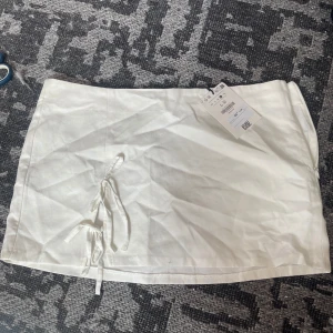 Vit kortkjol med knyt från Bershka - Superfräsch vit kortkjol från Bershka i linne- och bomullsblandning. Kjolen har snygga knytband framtill och dragkedja i sidan för enkel påklädning. Perfekt för en clean och trendig look i sommar. Behöver strykas, det är allt.