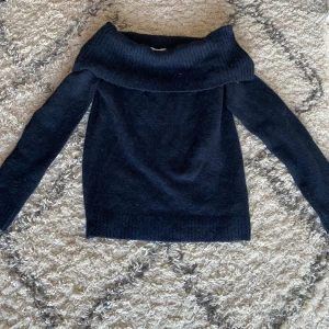 Mörkblå  offshoulder stickad tröja - Mysig svart stickad tröja med bred offshoulder-krage. Tröjan har lång ärm och ribbade muddar vid ärmslut och nederkant. 