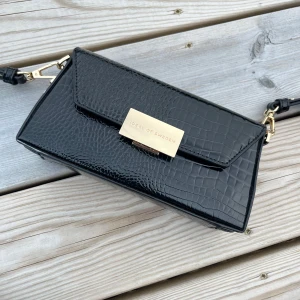 Svart axelväska från Ideal of Sweden Ny - Snygg svart axelväska från Ideal of Sweden med croco-mönstrad yta och guldfärgade detaljer. Väskan har en stilren, rektangulär form och justerbar axelrem. Perfekt för dig som vill ha en trendig och elegant accessoar med plats för det viktigaste. Ny