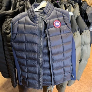 Mörkblå dunväst från Canada Goose - Snygg mörkblå quiltad dunväst från Canada Goose, modell Hybridge Lite Tech Vest. Västen har dragkedja framtill, ståkrage och klassisk logopatch på bröstet. Tillverkad i nylon och polyester med fyllning av gåsdun. Perfekt lager-på-lager-plagg för kyliga dagar.