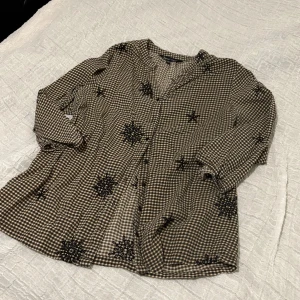 Rutig skjorta från Capsule med broderi - Snygg rutig skjorta från Capsule i beige och svart med unika svarta broderade detaljer. Skjortan har knappar framtill, lång ärm och en loose passform. Perfekt för dig som gillar en avslappnad men ändå cool stil.