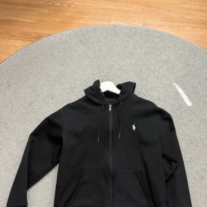 Svart hoodie från Polo Ralph Lauren - Svart hoodie från Polo Ralph Lauren med dragkedja framtill, huva med snörning och klassisk broderad logga på bröstet. Tröjan har känguruficka och är tillverkad i mjuk bomullsmix. Perfekt för en chill och stilren look. SNABB AFFÄR = BÄTTRE PRIS
