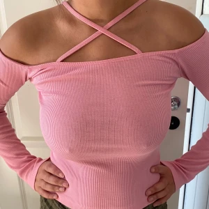 Ribbad rosa offshoulder topp - Säljer en ribbad rosa topp med offshoulder-design och långa ärmar. Toppen har smala axelband som korsas framtill för en snygg detalj. Figurnära passform och stretchigt material som sitter skönt. Perfekt för dig som gillar trendiga och feminina plagg.