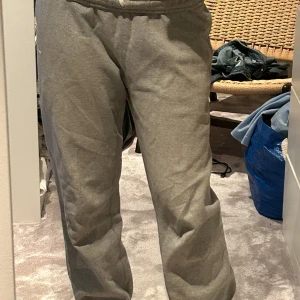 Gråa mjukisbyxor från Nike - Säljer ett par gråa mjukisbyxor från Nike med klassisk vit swoosh-logga på benet. Byxorna har resår i midjan med snörning och är tillverkade i mjuk bomullsblandning. Perfekta för chill eller träning.