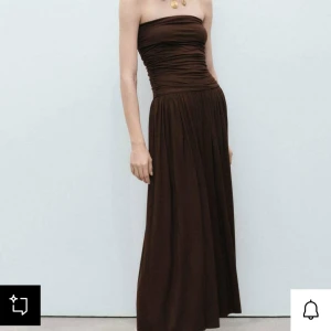 Brun axelbandslös långklänning - Säljer en snygg brun axelbandslös långklänning med rynkad överdel och flowy kjol. Perfekt för dig som gillar en stilren och elegant look. Klänningen har en tight passform upptill och faller snyggt från midjan och ner.