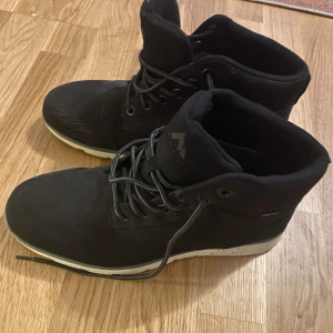 Svarta McKinley kängor strl 41 - Svarta kängor från McKinley, modell Richard AQX. Snygg design med högt skaft, snörning och vit sula. Tillverkade i syntetmaterial och passar perfekt för höst och vinter. Skorna har rund tå och är riktigt bekväma för vardagsbruk.