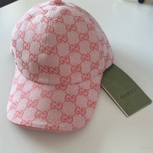 Rosa Gucci keps  - Snygg rosa keps från Gucci med ikoniskt GG-mönster i mörkare rosa över hela kepsen. Kepsen är tillverkad i bomull och har en klassisk böjd skärm samt en liten guldfärgad GG-detalj på sidan. Perfekt för dig som vill sticka ut med en lyxig accessoar.