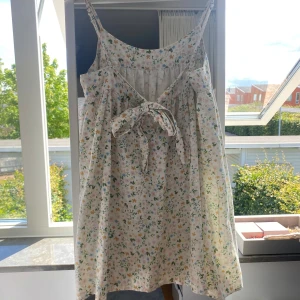 Blommig kort sommarklänning med rosett - Supersöt kort klänning med tunna axelband och blommigt mönster i pastellfärger. Lätt och luftig modell med v-ringning fram och öppen rygg med knytrosett. Superfint skick! Säljer för att den är för kort på mig. Nypris 1000kr.