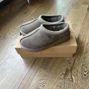 Grå mockatofflor från UGG - Mysiga grå tofflor från UGG i mocka med fluffigt foder och dekorativt mönster runt öppningen. Perfekta för kalla dagar, med platt sula och rund tå. Klassisk UGG-logga på sidan och robust yttersula.