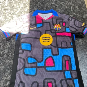 FC Barcelona Lamine Yamal 19 Nike tröja - Unik FC Barcelona fotbollströja med Lamine Yamal 19-tryck. Tröjan har ett grafiskt mönster i svart, blått, rosa och gult, med klubbmärke, Nike-logga och Spotify-sponsor framtill. Korta ärmar och blå krage. Materialet är lätt och andas bra.