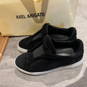 Svarta sneakers från Axel Arigato - Stilrena svarta sneakers från Axel Arigato i mocka med vit platt sula. Skorna har minimalistisk design utan synlig snörning och en diskret logga på plösen. Perfekta för dig som gillar en clean och modern look.