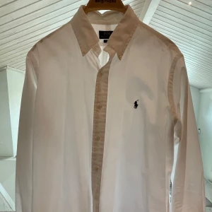 Vit skjorta från Polo Ralph Lauren  - Klassisk vit skjorta från Polo Ralph Lauren i bomull, med broderad marinblå logga på bröstet. Skjortan har lång ärm, knappar framtill och button down-krage. Passformen är normal (classic fit) och materialet är mjukt och skönt.