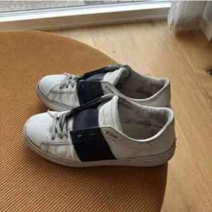 Valentino Open sneakers vit/blå - Snygga Valentino Open sneakers i vitt skinn med bred mörkblå rem över vristen. Klassisk rund tå och platt sula, perfekt för en clean och trendig look. Diskret Valentino-logga på plösen och stilrena detaljer.