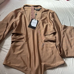 Beige satinblus med cut outs från NAANAA - Snygg beige blus från NAANAA i satinliknande material. Modellen har långa ärmar, djup v-ringning och coola cut outs på sidorna som ger en edgy vibe. Perfekt för dig som vill sticka ut med en trendig och unik topp.