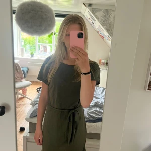 Olivgrön klänning från Bik Bok - Olivgrön kortklänning från Bik Bok i mjuk bomull. Klänningen har korta ärmar, rund hals och ett matchande knytbälte i midjan. Öglan som knytbältwr ska dras igenom är trasigt på ena sidan, men annars inga deffekter.Säljer då den inte längre kommer till användning.