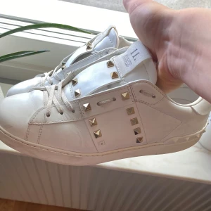 Valentino open - Sjukt snygga vita Valentino Rockstud-sneakers i skinn med de ikoniska guldiga nitarna längs sidorna och på hälen. Klassisk rund tå, snörning framtill och en platt sula som är superbekväm. Perfekta för dig som vill addera en lyxig och edgy vibe till vilken outfit som helst. Två små spikar saknas men inget annat att anmärka på, så tveka inte att höra av dig om du har frågor eller funderingar 🙌🤝💠💎 Box eller kvitto finns tyvärr inte men skorna är 100% äkta