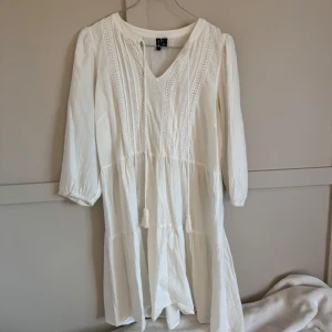 Vit långärmad klänning från Vero Moda - Superfin vit kortklänning från Vero Moda med v-ringning, knytband med tofsar och spetsdetaljer framtill. Klänningen har trekvartsärm och är tillverkad i ett lätt bomullstyg som känns luftigt och skönt. Perfekt för varma dagar och en avslappnad stil.