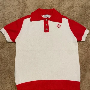 Rödvit pikétröja Casablanca Tennis Club - Snygg pikétröja från Casablanca Tennis Club i rött och vitt. Tröjan har korta ärmar, pärlknappar och broderad logga på bröstet. Klassisk krage och ribbade muddar. Perfekt för en sportig och trendig look.