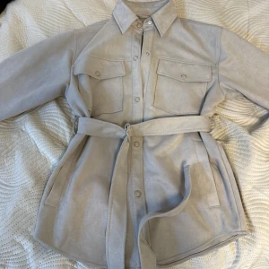 Beige overshirt från Pull&Bear - Snygg beige overshirt från Pull&Bear med bälte i midjan och stora bröstfickor. Jackan har knappar framtill och är i ett mjukt mockaliknande material. Perfekt för lager-på-lager och ger en chill men stilren vibe.