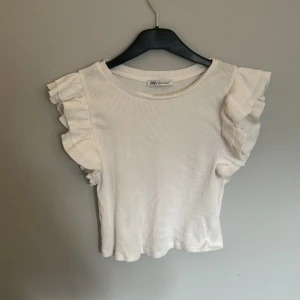 Vit ribbad topp med volangärm Zara - Supersöt vit ribbad topp från Zara med volangärmar som ger en extra touch. Modellen är croppad och har rund halsringning. Perfekt för dig som gillar en feminin och trendig stil. Materialet är mjukt och stretchigt, vilket gör den riktigt skön att bära.