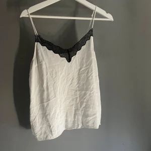 Vit linnetopp med spets från H&M - Säljer en vit linnetopp från H&M med tunna axelband och snygg svart spetsdetalj längs v-ringningen. Toppen är i ett lätt och svalt material, perfekt för varma dagar eller lager på lager. Enkel men ändå chic stil som lyfter vilken outfit som helst.