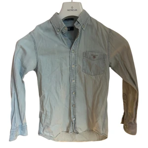 Ljusblå skjorta från Gant i bomull - Klassisk ljusblå skjorta från Gant med button down-krage och bröstficka med broderad logga. 134-140cm för 9-10år bra skick men två små färgfläckar på framsidan som knappt syns. Skjortan har långa ärmar och knäppning framtill. Tillverkad i mjuk bomull, perfekt för en avslappnad men ändå stilren look.