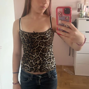 Leopardmönstrad linne med rosett - Säljer ett leopardmönstrat linne med tunna svarta axelband och en söt liten rosett framtill. Toppen har svarta spetsdetaljer längs kanten och är figurnära. Perfekt för dig som vill sticka ut med ett djurmönster och cool vibe.