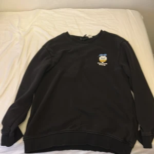 Svart Disneytröja med Kalle Anka-tryck - Svart sweatshirt från Disney med broderad Kalle Anka på bröstet och stort tryck av Knatte, Fnatte och Tjatte på ryggen. Tröjan har rund hals, långa ärmar och är gjord i mjuk bomullsmix.