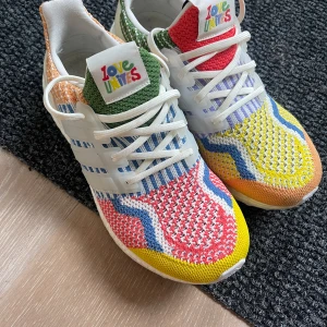 Adidas Ultraboost Love Unites sneakers 5.0 - Färgglada Adidas Ultraboost Love Unites sneakers med stickad ovandel i rött, gult, grönt, blått och orange. Skorna har vita snören, vit sula och broderad Love Unites-logga på plösen. Mönstrad design med vågiga detaljer och klassiska tre ränder på sidan.