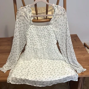 Vit blommig kortklänning Vero Moda - Supersöt vit kortklänning från Vero Moda med små gröna blommor, smockad överdel och långa, transparenta ärmar med volang vid ärmslut. Klänningen har en luftig känsla och är perfekt för varma dagar. Romantisk och trendig look!