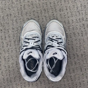 Grå Asics GEL sneakers med mesh - Snygga Asics GEL sneakers i grått med silvriga detaljer och svart logga. Ovandelen är i mesh och syntet för en sportig vibe. Skorna har snörning och en chunky sula som ger extra komfort. Perfekta för dig som gillar streetstyle och sportig look.