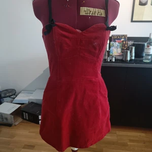 Röd klänning med svarta band - Supersöt röd kortklänning med svarta axelband och rosetter. Figurnära passform och hjärtformad urringning framtill. Dragkedja i ryggen för enkel påklädning. Perfekt för dig som vill sticka ut med en retrovibe!