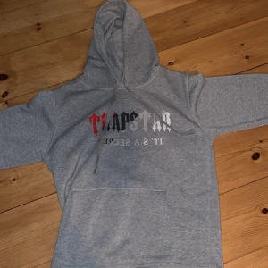 Grå Trapstar hoodie med tryck - Tja,Säljer en grå hoodie från Trapstar med fet logga i svart, rött och vitt framtill. Klassisk känguruficka, huva med snörning och mjukt material. Perfekt för dig som gillar streetwear och vill sticka ut. Om ni har några frågor så är det bara att skriva.