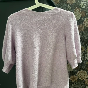 Ljuslila stickad tröja Vero Moda - Supermjuk ljuslila stickad tröja från Vero Moda med korta puffärmar och ribbade muddar. Perfekt för dig som gillar en söt och trendig look. Tröjan har en rund halsringning och är i en skön, lite fluffig kvalitet. Lite nopprig som man ser på bilden plus att den är mer lila i verkligenhegen än på bilden💜