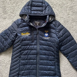 blå jacka  - Mörkblå pufferjacka från svenska ridsportförbundet  med huva och broderade loggor för Sweden Supporter och Svenska Ridsportförbundet. Jackan har dragkedja framtill, quiltad design och två sidofickor. 