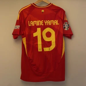 Spaniens Lamine Yamal matchtröja #19 - Säljer en röd Spanien fotbollströja replika från Adidas med gula detaljer, nummer 19 och Lamine Yamal på ryggen. Tröjan har korta ärmar, broderat landslagsmärke, Nations League-märke och gula ränder på axlarna. Tillverkad i lätt och ventilerande material. Tvättas för hand!