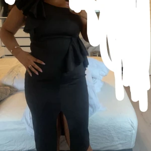 Svart oneshoulder klänning med volang - Supersnygg svart långklänning från Sasha med oneshoulder och stora volangdetaljer vid axel och höft. Klänningen har en slits framtill och figurnära passform som ger en elegant siluett. Perfekt för dig som vill sticka ut med stilren look.