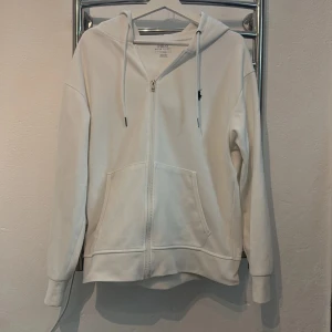 Vit hoodie med dragkedja från Polo Ralph Lauren - Vit hoodie från Polo Ralph Lauren med dragkedja framtill och klassisk logga broderad på bröstet. Tröjan har huva med dragsko och en stor magficka. Perfekt för en avslappnad stil. Hoodien är i toppskick 10av10!!
