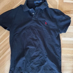 Svart pikétröja från Polo Ralph Lauren - Klassisk svart pikétröja från Polo Ralph Lauren med korta ärmar och röd broderad logga på bröstet. Tröjan har krage och knäppning med två knappar. Perfekt för en stilren och avslappnad look.