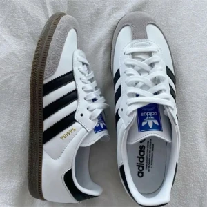 Adidas Samba - Klassiska Adidas Samba sneakers i vitt skinn med svarta ränder och grå mockadetaljer på tån. Snygg gummisula och ikonisk blå Adidas-logga på plösen. Perfekta för en stilren och sportig look.