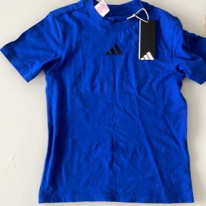 Blå Adidas t-shirt i bomull - Snygg blå t-shirt från Adidas med klassisk logga på bröstet. T-shirten är i mjuk bomull och har en enkel, sportig look med rund hals och korta ärmar. Perfekt för dig som gillar stilren och bekväm streetwear.