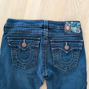 True Religion bootcut jeans blå strl 27 - Säljer nu mina sjukt snygga, vintage och unika true religon jeans med dekorativa kristaller/stenar på knapparna i storlek 27🥰   Några av stenarna på jeansknappen fram saknas och jeansen är lite söndertrampade men inget som stör användningen! Midjemått: 37 cm rakt över Innerbenslängd: 76 cm  Pris kan diskuteras!