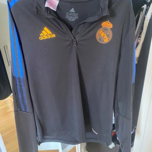 Real Madrid långärmad träningströja Adidas - Snygg svart långärmad träningströja från Adidas med Real Madrid-logga och orange detaljer. Tröjan har halv dragkedja, blå ränder på ärmarna och är tillverkad i funktionsmaterial. Perfekt för fotbollsträning eller chill.