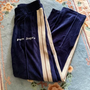 Palm Angels blå mjukisbyxor L - Mörkblå mjukisbyxor från Palm Angels med beige sidoränder och broderad logga på vänster ben. Byxorna har resår i midjan och är gjorda i mjuk polyester med en skön loose passform. Perfekta för en chill och trendig streetwear-look.