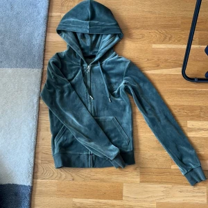 Grön velour hoodiejacka från Juicy Couture - Mjuk och lyxig grön hoodiejacka i velour från Juicy Couture. Jackan har dragkedja, huva med snörning och broderad JC-logga på bröstet. Perfekt för en chill och trendig look.