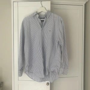 Randig skjorta från Ralph Lauren - Buda gärna! Klassisk skjorta från Ralph Lauren i vitt och blått med tunna ränder. Skjortan har lång ärm, knappar framtill och broderad logga på bröstet. Tillverkad i mjuk bomull och har en tidlös, normal passform. Perfekt för en clean och stilren look. Stlk 10 vilket motsvarar Large eller EU 40. Perfekt skick!